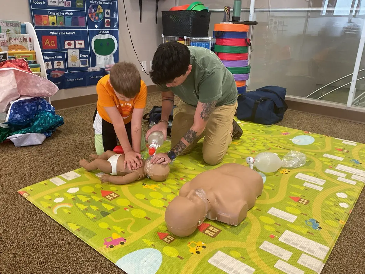 An Atlas instructor guiding a young learner through infant CPR on a training manikin, with an adult manikin and bag-valve masks nearby
