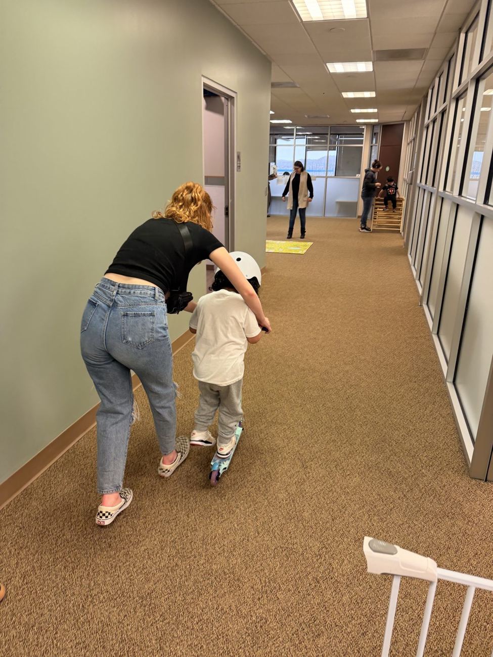 A therapist helping a child on a scooter in a bright hallway