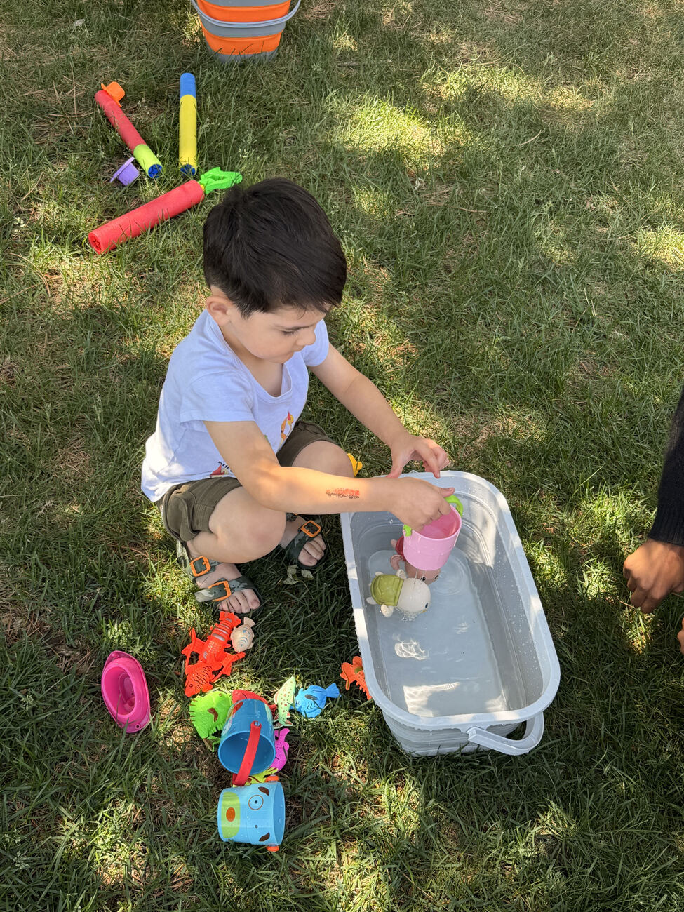 A young child playing with cups and toys in a tub of water on the grass