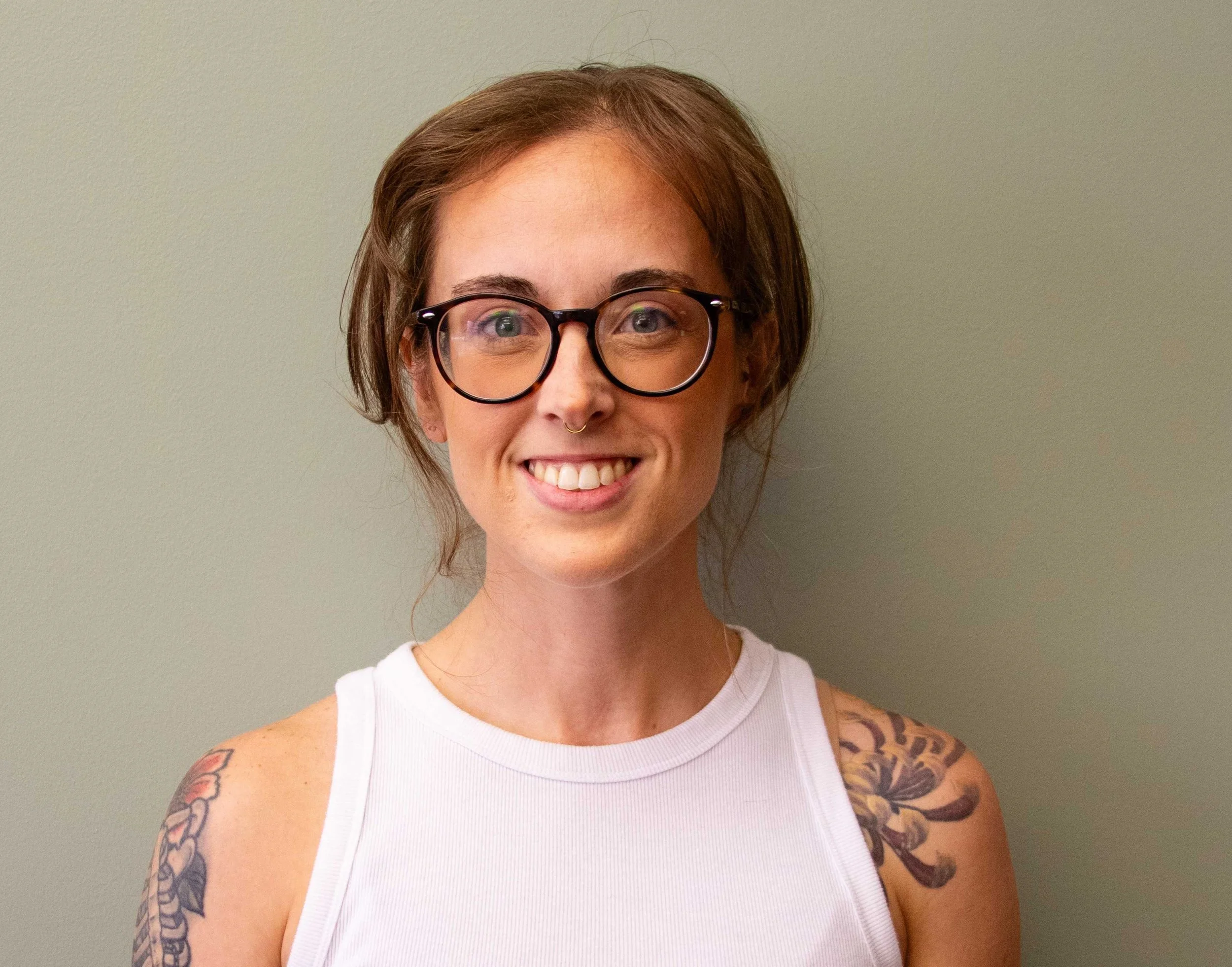 Sarah Anna, smiling, wearing round glasses and a white tank top in front of a sage green wall