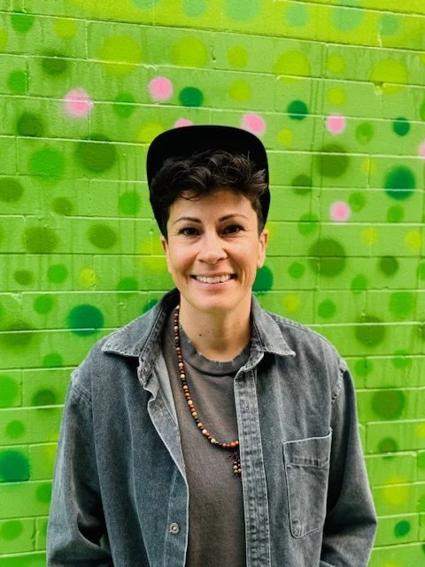 Tori Salas, smiling, wearing a black cap and denim jacket in front of a green painted brick wall